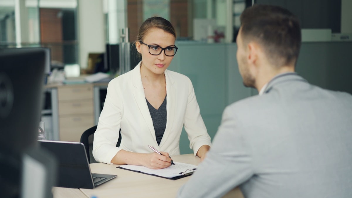 Friendly young businesswoman in glasses and suit is interviewing a male candidate for job in office. People are talking asking and answering questions, girl is writing.